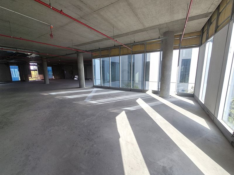 An expansive, unfinished open-plan office space featuring exposed concrete ceilings, structural columns, and large floor-to-ceiling windows providing ample natural light.