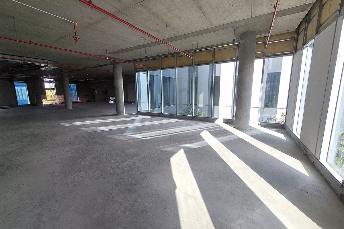An expansive, unfinished open-plan office space featuring exposed concrete ceilings, structural columns, and large floor-to-ceiling windows providing ample natural light.