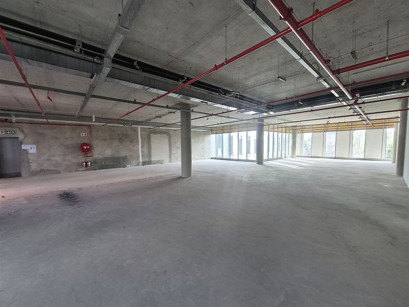 A large, open-plan office space in shell condition featuring exposed concrete ceilings, fire suppression systems, and expansive floor-to-ceiling windows.