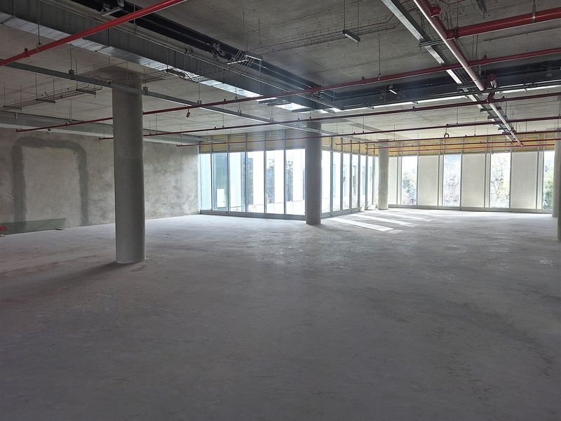 A large, open-plan office space in shell condition, featuring exposed concrete ceilings, fire sprinkler systems, and expansive floor-to-ceiling windows providing natural light.