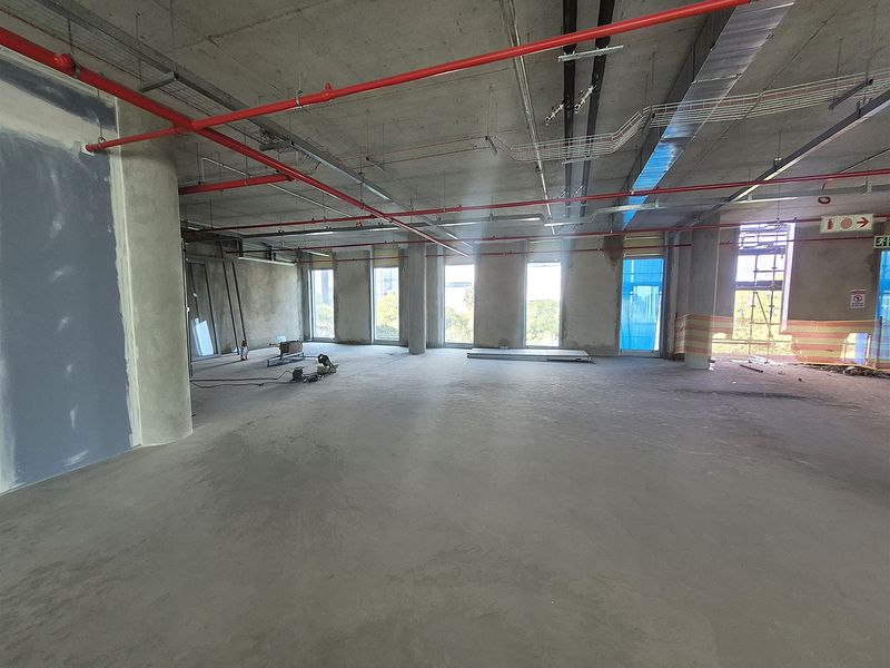 An expansive, open-plan office space currently in a shell condition, featuring exposed industrial ceilings, concrete flooring, and large windows providing natural light.