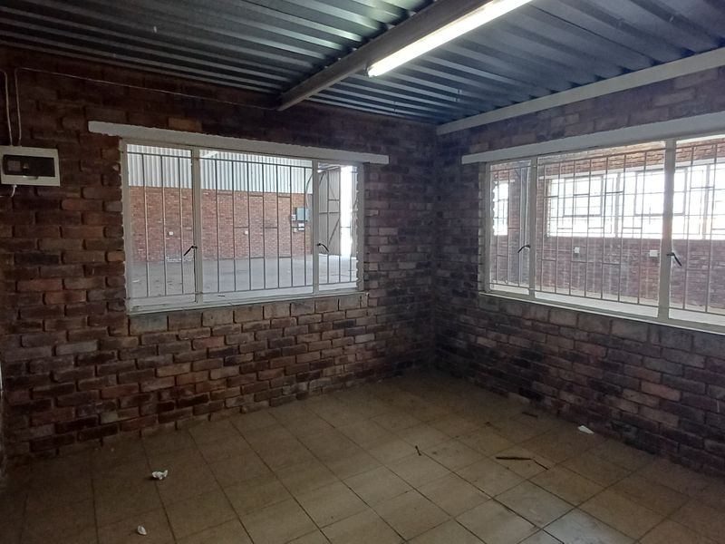An empty industrial office space featuring exposed brick walls, tiled flooring, and a corrugated metal ceiling with security-barred windows.