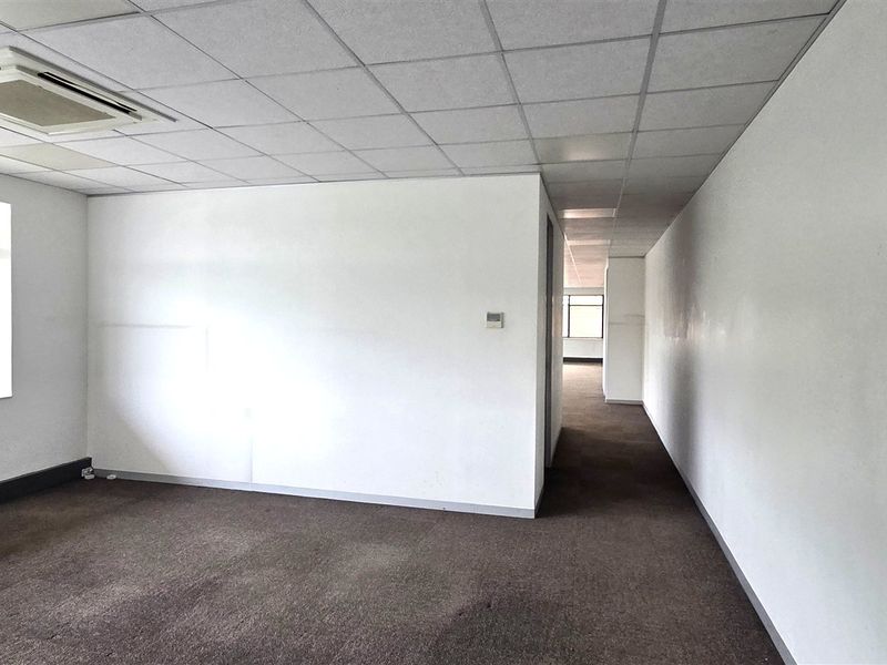 An empty, open-plan office space featuring a suspended ceiling, recessed air conditioning, and carpeted flooring with large windows providing natural light.