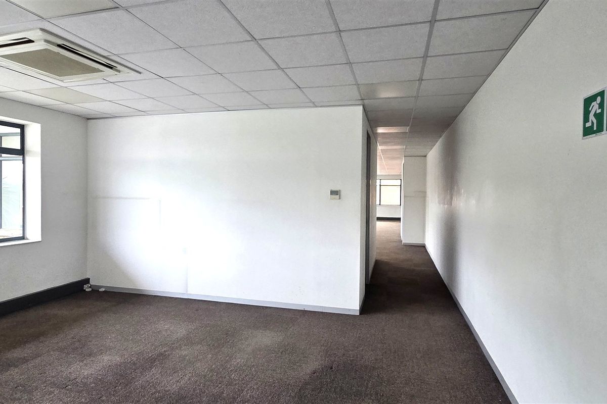 An empty, open-plan office space featuring a suspended ceiling, recessed air conditioning, and carpeted flooring with large windows providing natural light.