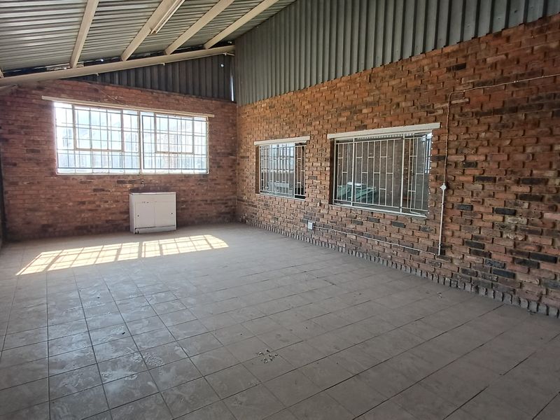 An empty industrial utility space featuring exposed brick walls, tiled flooring, and large windows with security bars under a metal roof structure.