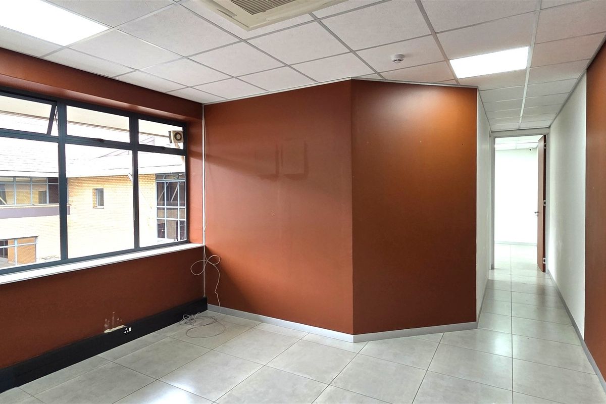 A bright, empty office space featuring tiled flooring, a suspended ceiling with recessed lighting, and large windows providing natural light.