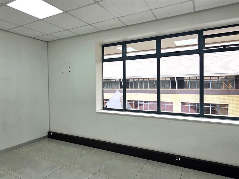 A vacant, well-lit office space featuring tiled flooring, a suspended ceiling, and large windows providing natural light and exterior views.