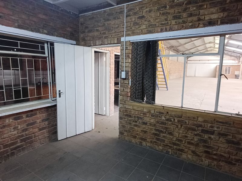 An empty utility or office space featuring exposed brick walls, tiled flooring, and large windows looking into the main industrial warehouse area.