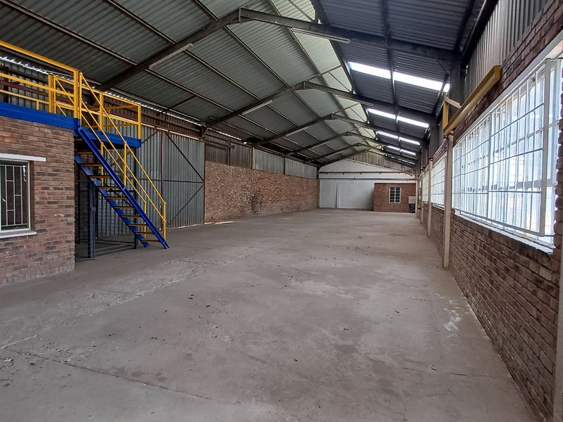 Spacious industrial warehouse featuring exposed brick walls, concrete flooring, steel roof trusses, and a mezzanine level with access via a steel staircase.