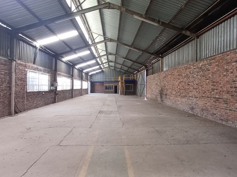 Spacious industrial warehouse featuring exposed brick walls, high steel truss ceilings with skylights, concrete flooring, and a mezzanine level for additional storage or office space.