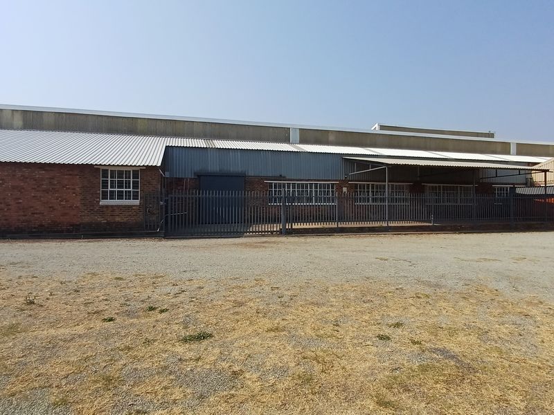 Front elevation of an industrial warehouse featuring brick construction, corrugated metal roofing, and secure steel perimeter fencing with industrial-style windows.