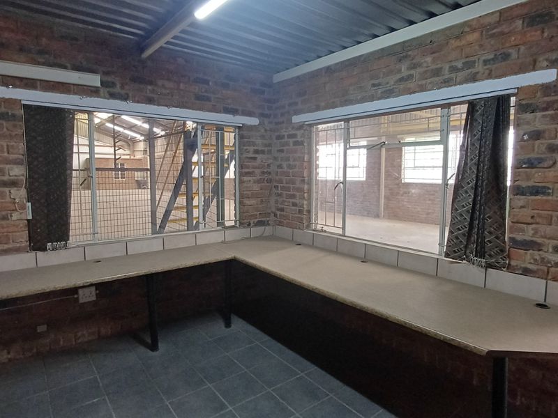 Industrial-style office space featuring exposed brick walls, a built-in wrap-around desk, tiled flooring, and large windows overlooking the warehouse area.