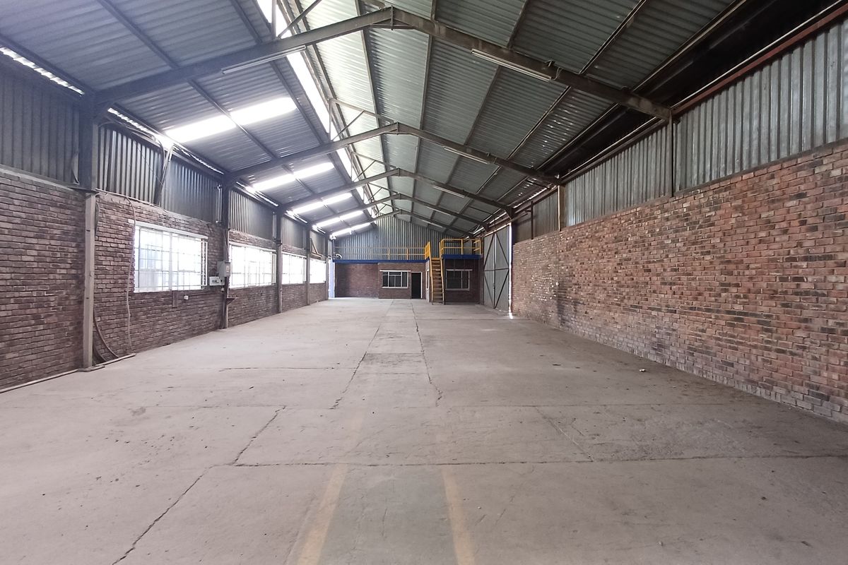 Spacious industrial warehouse featuring exposed brick walls, high steel truss ceilings with skylights, concrete flooring, and a mezzanine level for additional storage or office space.