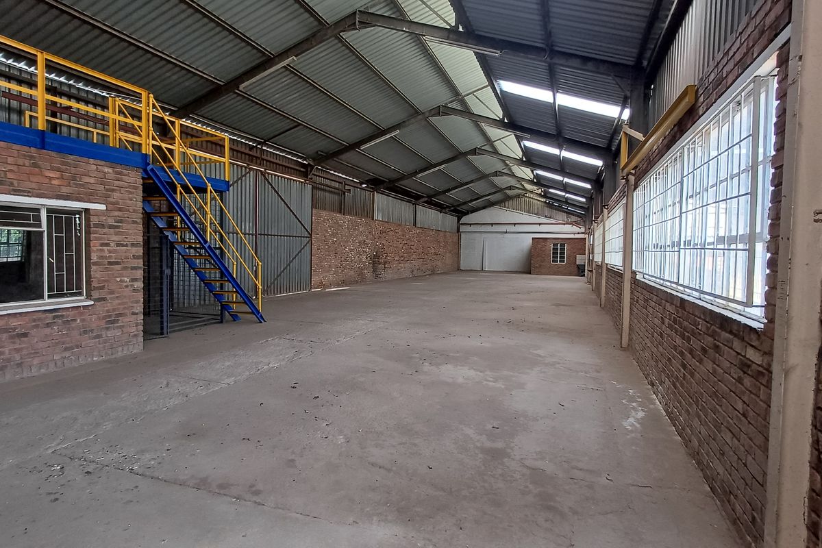 Spacious industrial warehouse featuring exposed brick walls, concrete flooring, steel roof trusses, and a mezzanine level with access via a steel staircase.