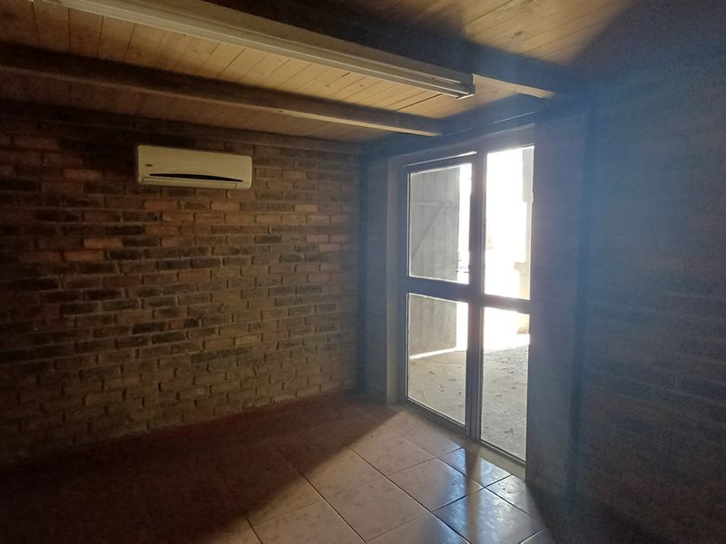 An empty office space featuring exposed brick walls, timber ceiling, tiled flooring, and a split-system air conditioner with direct access to an outdoor area.