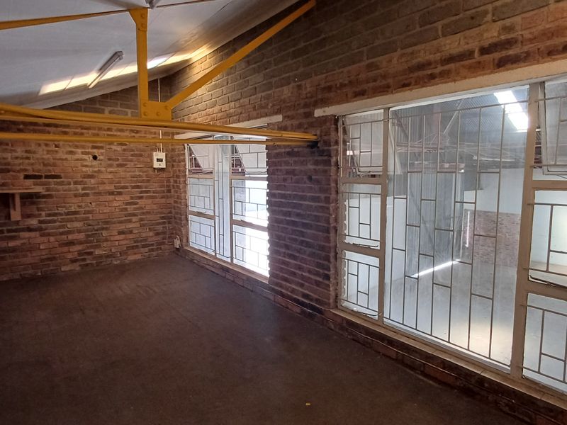 Industrial utility space featuring exposed brick walls, steel roof trusses, and large steel-framed windows with security bars, including a basic built-in sink unit.
