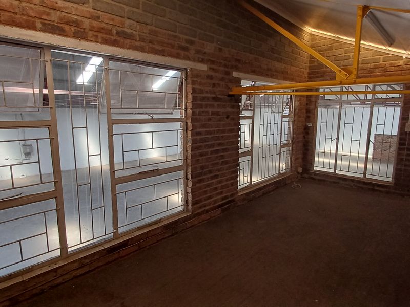 Spacious industrial open-plan area featuring exposed brick walls, steel roof trusses, and large windows with security bars, offering a versatile blank canvas.