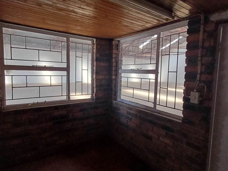 Small office space featuring exposed brick walls, timber ceiling, and security-barred windows overlooking the main industrial warehouse area.