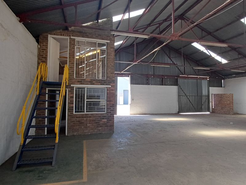 Spacious industrial warehouse featuring high ceilings with exposed steel trusses, concrete flooring, and a built-in brick mezzanine office structure with staircase access.