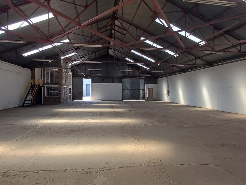 Spacious industrial warehouse featuring high exposed steel trusses, concrete flooring, natural light from skylights, and an integrated mezzanine office area.