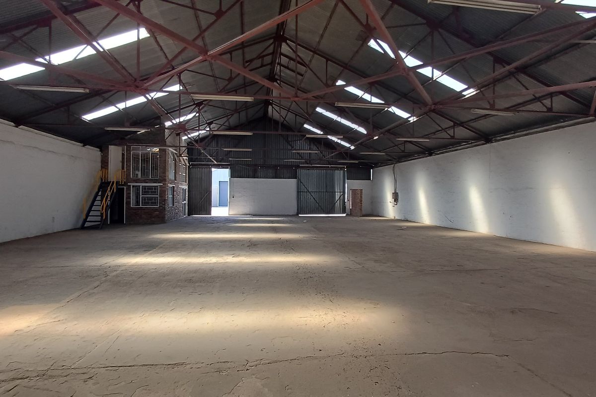 Spacious industrial warehouse featuring high exposed steel trusses, concrete flooring, natural light from skylights, and an integrated mezzanine office area.