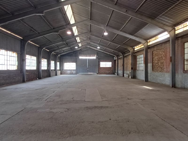 Spacious industrial warehouse featuring a high-clearance steel portal frame, concrete flooring, exposed brick walls, and large steel-framed windows providing ample natural light.