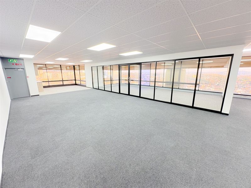 A spacious, modern open-plan office area featuring glass partitions, carpeted flooring, and a suspended ceiling with recessed lighting.