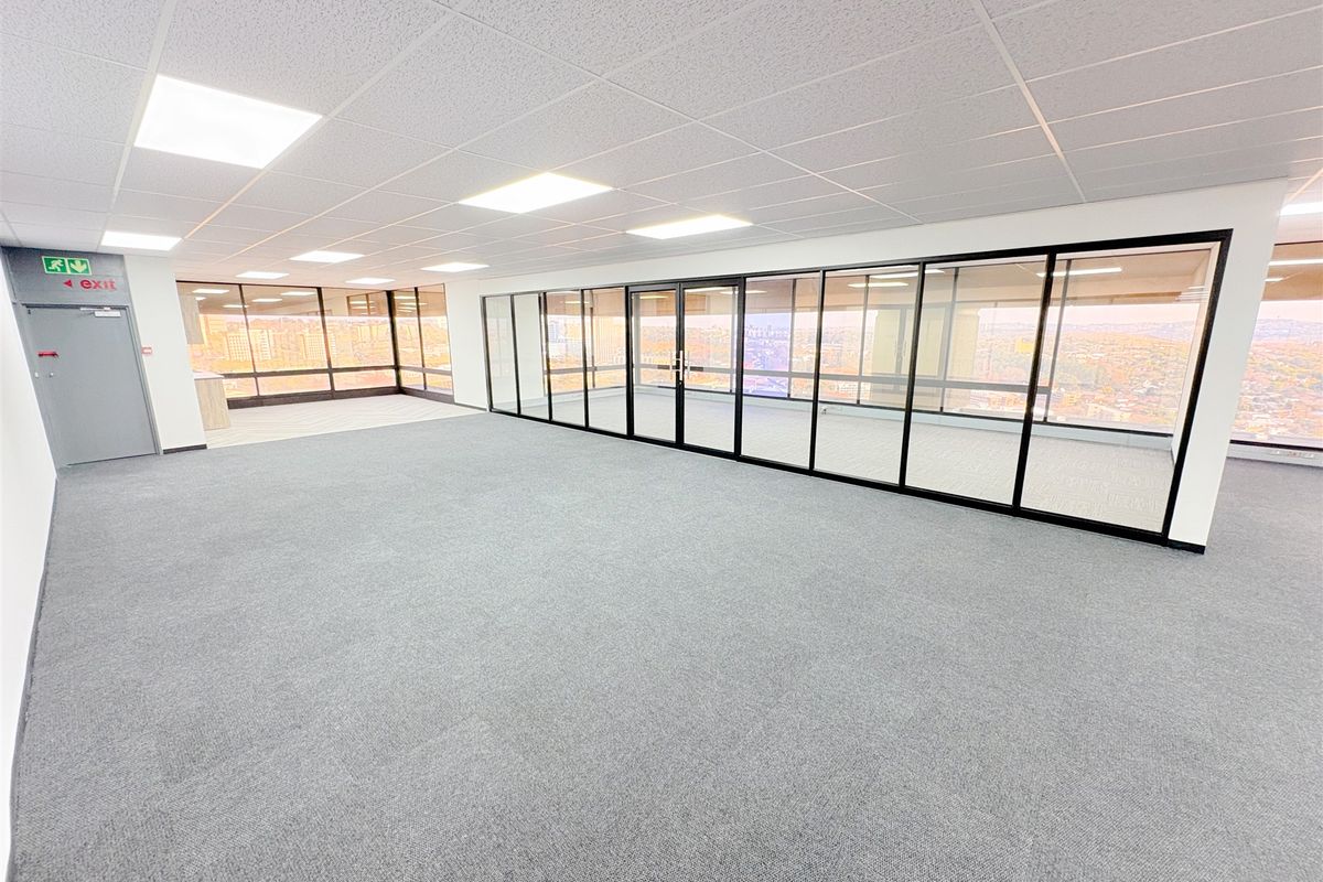 A spacious, modern open-plan office area featuring glass partitions, carpeted flooring, and a suspended ceiling with recessed lighting.