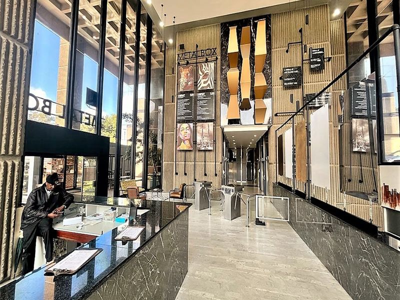 Modern office building entrance lobby featuring high ceilings, floor-to-ceiling glass windows, marble reception desk, and secure turnstile access points.