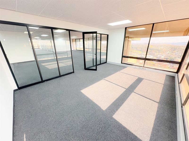 A bright, modern, and empty office space featuring glass partitions, carpeted flooring, and large windows offering expansive views.