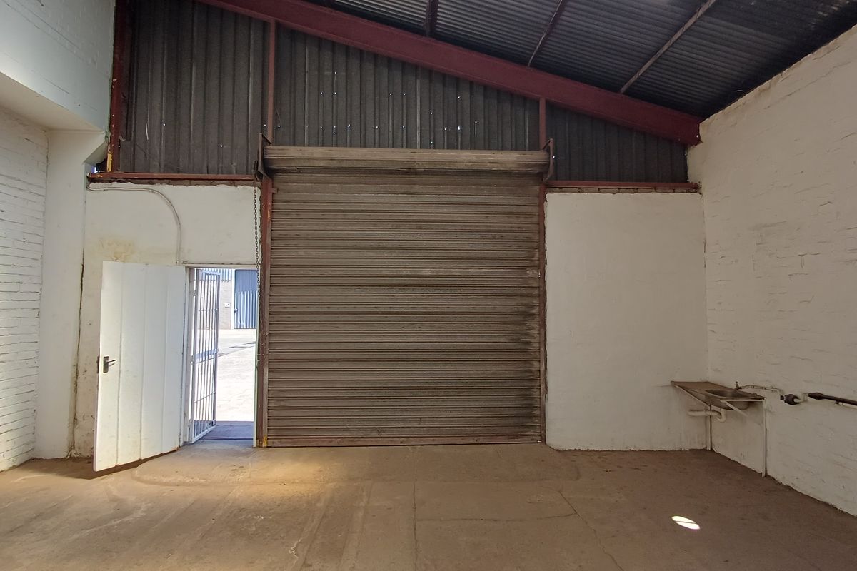 Industrial utility space featuring a large roller shutter door, exposed roof structure, painted brick walls, and a wall-mounted utility sink.