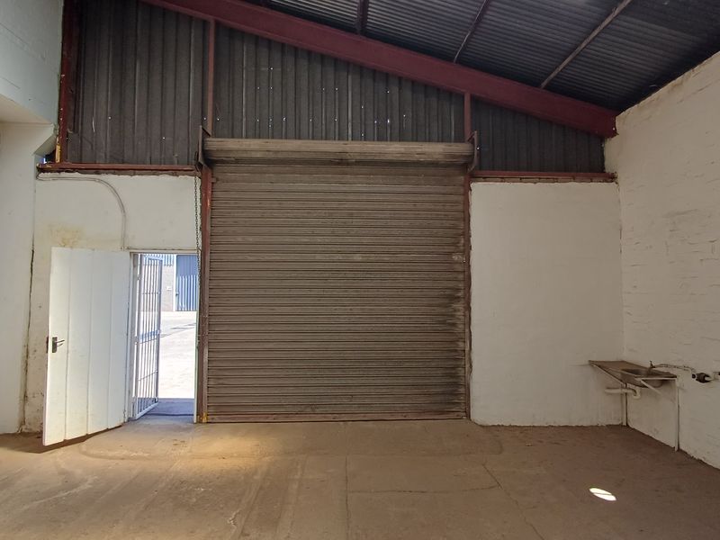 Industrial utility space featuring a large roller shutter door, exposed roof structure, painted brick walls, and a wall-mounted utility sink.