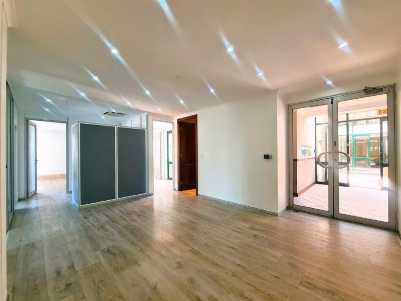A bright, open-plan office space featuring modern laminate flooring, recessed ceiling lighting, and glass double doors leading to an outdoor area.