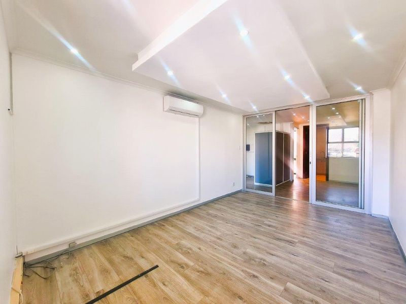 A bright, modern office space featuring laminate wood flooring, a recessed ceiling with downlights, a split-system air conditioner, and a sliding glass partition.