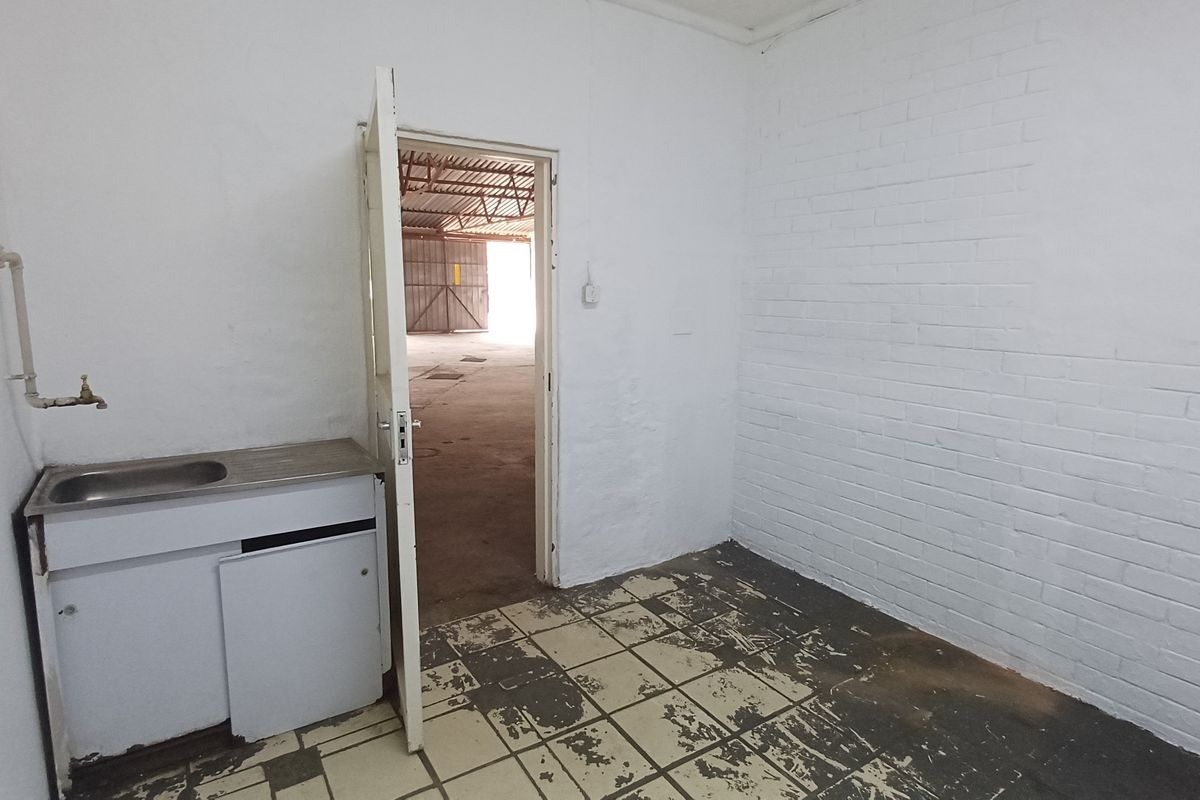 A small utility space featuring a stainless steel sink, painted brick walls, and worn tiled flooring, providing direct access to the main industrial warehouse area.