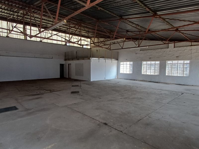 Spacious industrial utility area featuring high-volume exposed steel roof trusses, concrete flooring, and multiple security-barred windows providing ample natural light.
