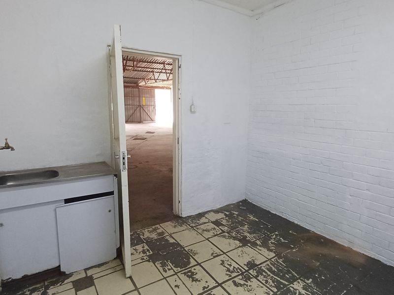 A small utility space featuring a stainless steel sink, painted brick walls, and worn tiled flooring, providing direct access to the main industrial warehouse area.
