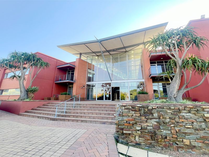 Modern office building exterior featuring a prominent glass facade, stone retaining walls, and a paved entrance area with architectural steel roofing.
