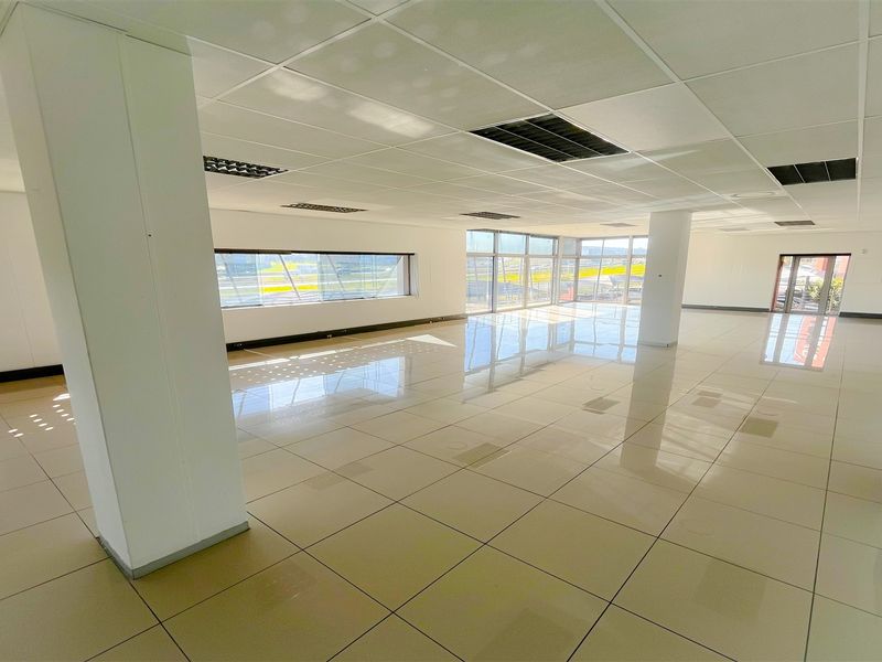 A spacious, open-plan office area featuring polished tiled flooring, a suspended ceiling with recessed lighting, and large windows providing ample natural light.