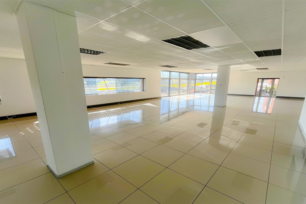 A spacious, open-plan office area featuring polished tiled flooring, a suspended ceiling with recessed lighting, and large windows providing ample natural light.