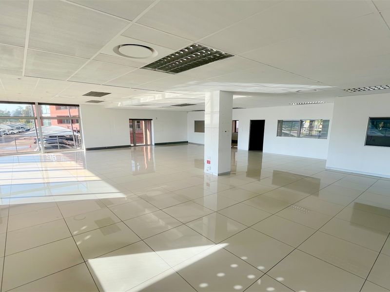 A spacious, open-plan office area featuring polished tiled floors, a suspended ceiling with recessed lighting, and large windows providing ample natural light.