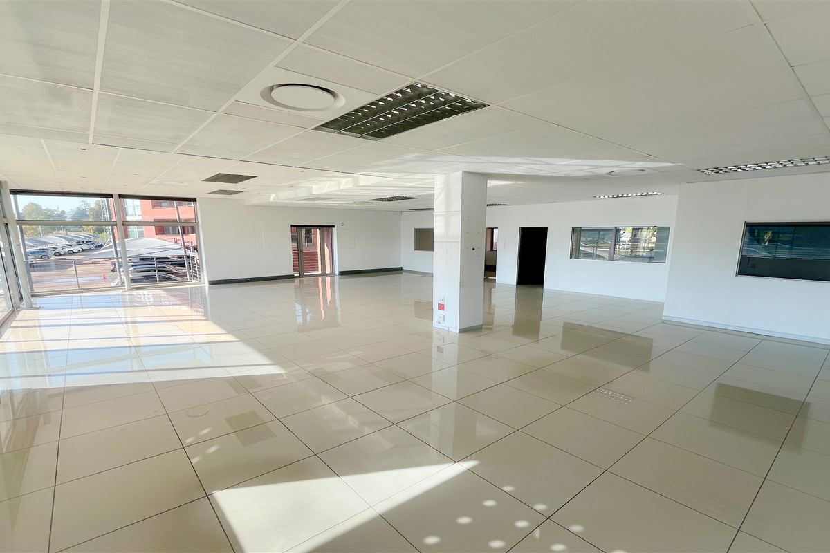 A spacious, open-plan office area featuring polished tiled floors, a suspended ceiling with recessed lighting, and large windows providing ample natural light.