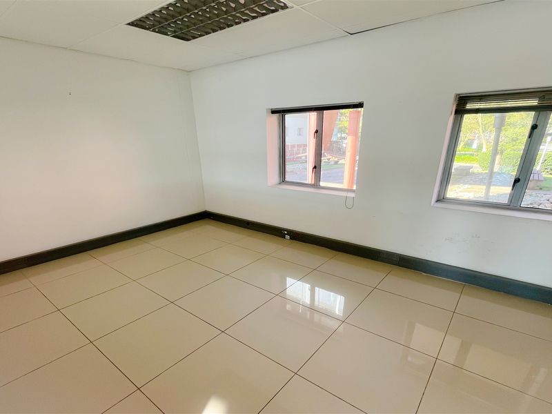 A bright, empty office space featuring neutral tiled flooring, recessed ceiling lighting, and large windows providing natural light.