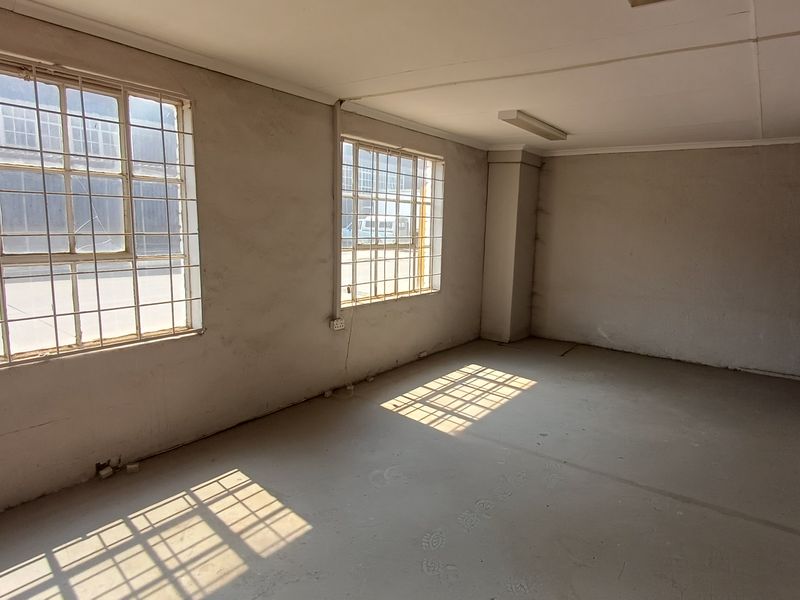 An empty, industrial-style office space featuring concrete floors, steel-framed windows with security bars, and basic fluorescent lighting, requiring cosmetic updates.