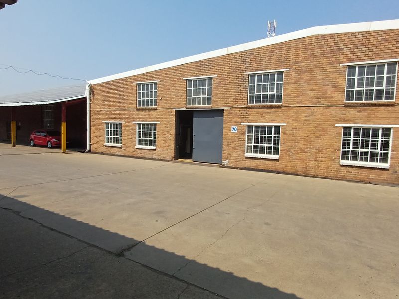 Industrial warehouse exterior featuring a brick facade, large sliding access door, steel-framed windows, and a spacious concrete yard with a covered loading area.