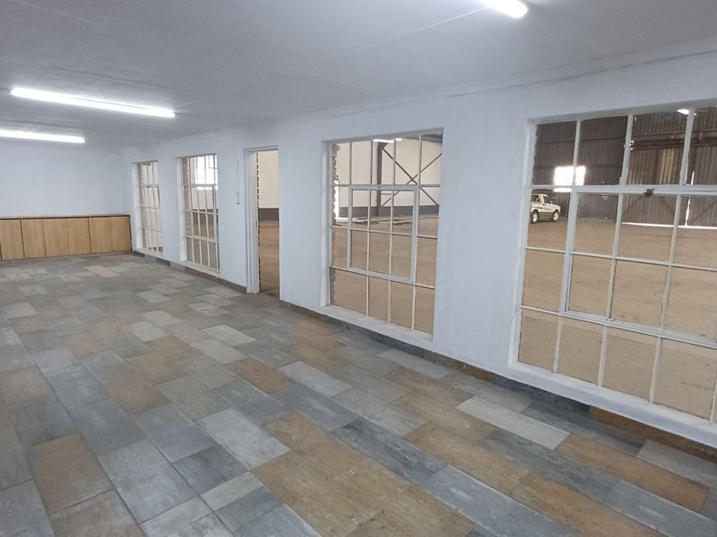 An open-plan office space featuring tiled flooring, built-in cabinetry, and large steel-framed windows overlooking an industrial warehouse area.