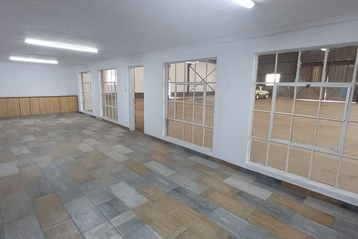 An open-plan office space featuring tiled flooring, built-in cabinetry, and large steel-framed windows overlooking an industrial warehouse area.