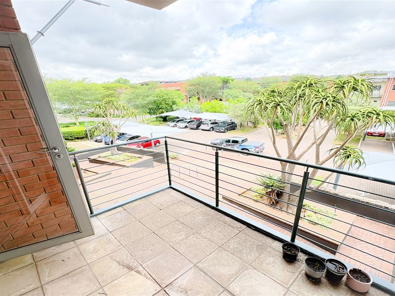 A private balcony featuring tiled flooring and a metal railing, overlooking the office complex parking area and landscaped grounds.