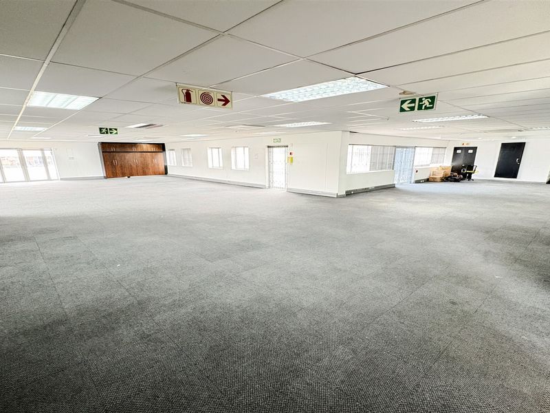 A spacious, open-plan office area featuring neutral carpet tiles, a suspended ceiling with recessed lighting, and integrated storage cabinetry.
