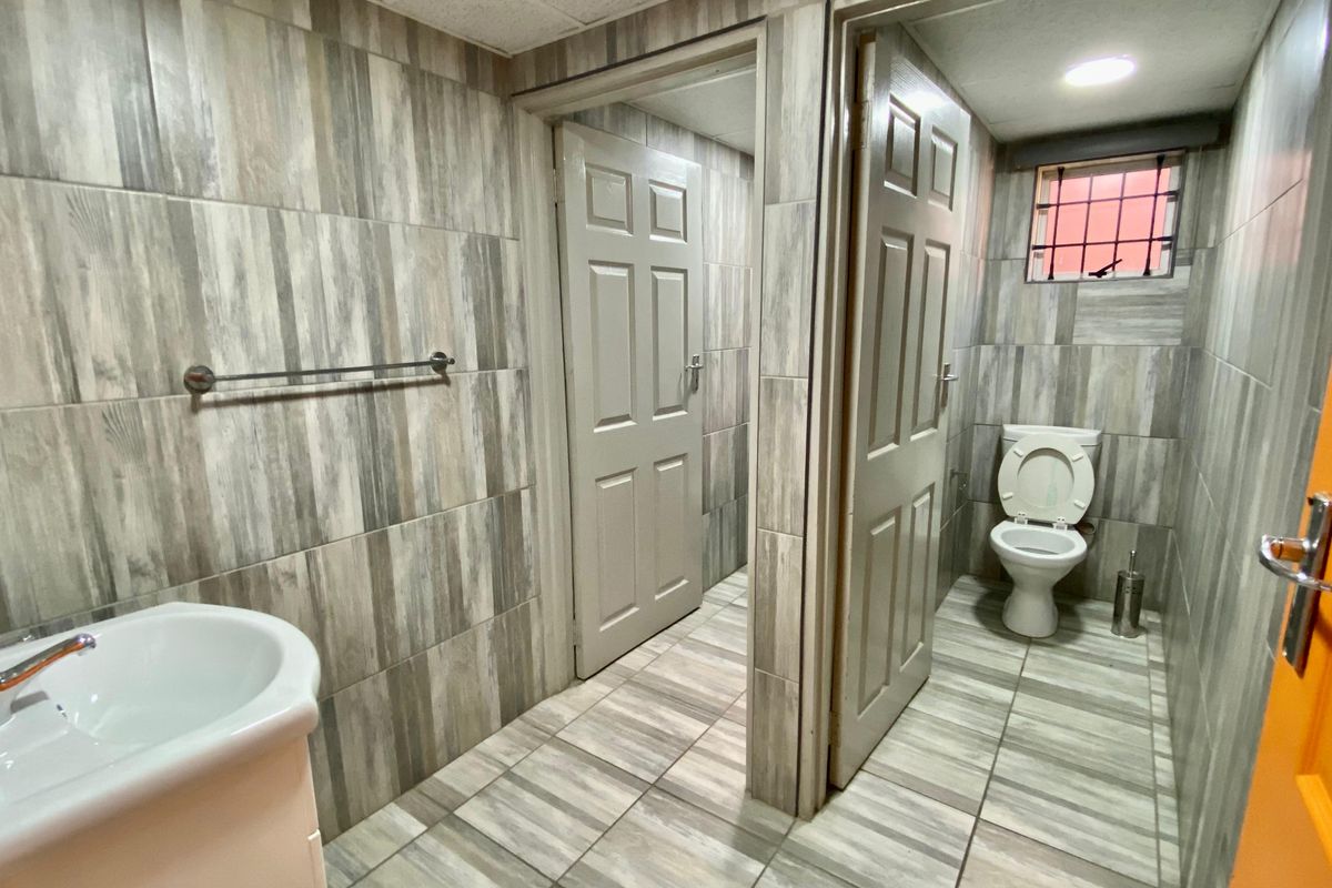 A clean, tiled commercial bathroom featuring a pedestal washbasin, a separate toilet cubicle, and modern wood-look ceramic wall and floor tiling.